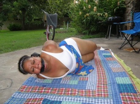 Mature ebony plumper is hogtied on a comforter while cleave gagged